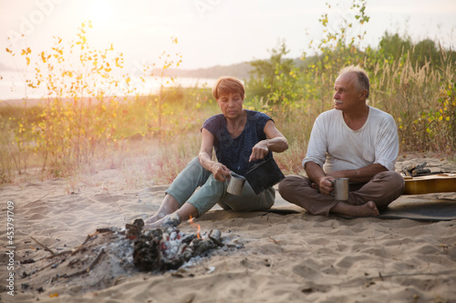 Senior couple Camping holiday in the summer nature and drink hot tea on the beach near the fire. Camping concept. 
