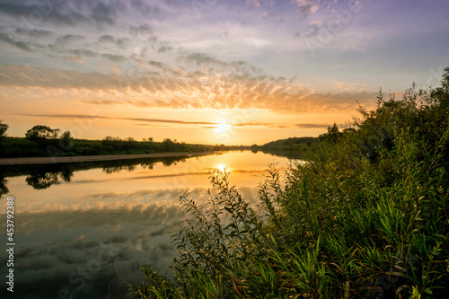 Scenic view at beautiful summer river sunset with reflection on water with green bushes  calm water  deep colorful cloudy sky and glow on horizon on a background  spring evening landscape