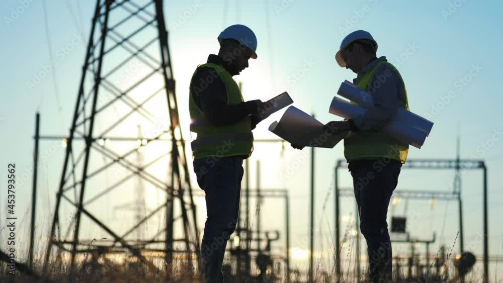 Teamwork industry a concept. Two engineers electric experts in protective helmets discussing the construction of a power line. Silhouette Of Engineers Standing On Field With Electricity Towers. Teamwork industry a concept. Two engineers electric experts in protective helmets discussing the construction of a power line. Silhouette Of Engineers Standing On Field With Electricity Towers.