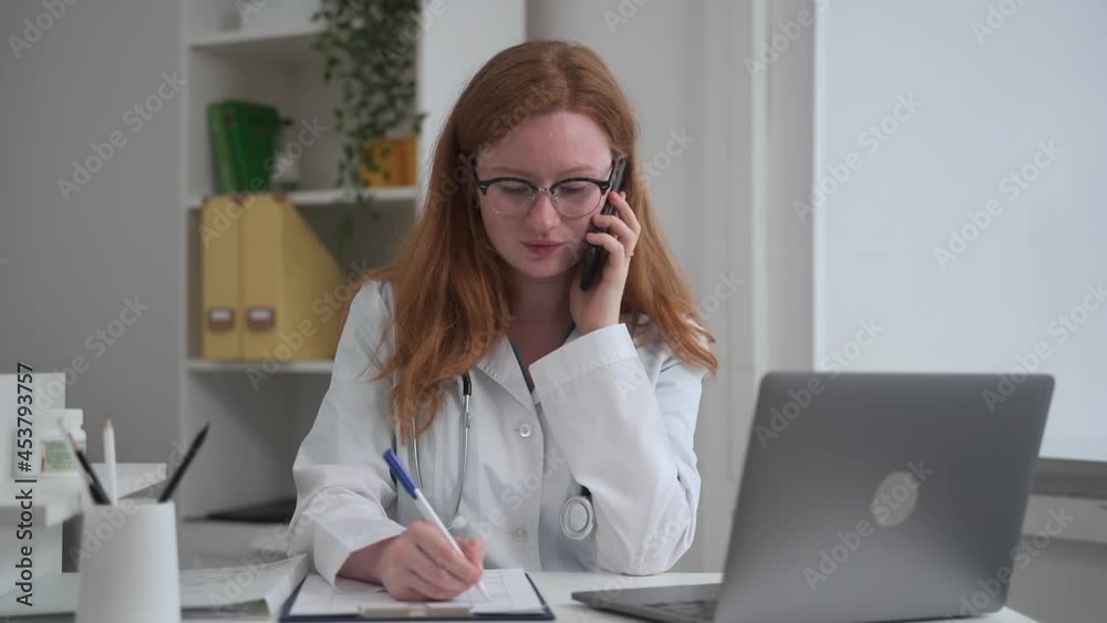Doctor ehealth online service. Spbas Positive redhead general practitioner woman with glasses consults client by phone writing at table in hospital office Doctor ehealth online service. Spbas Positive redhead general practitioner woman with glasses consults client by phone writing at table in hospital office
