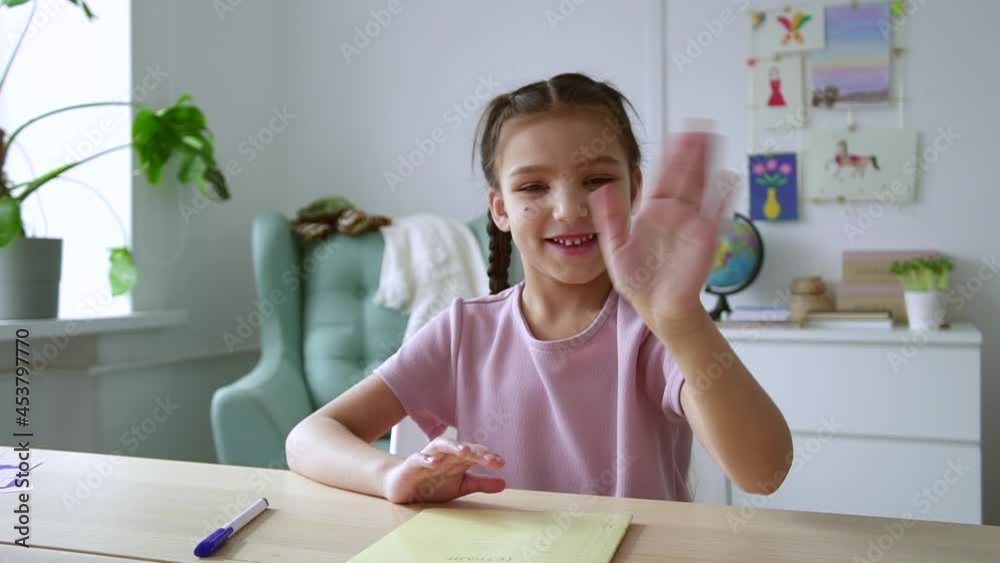 Online primary school education. Spbd Cheerful little girl in pink t-shirt waves hand to greet teacher and friends at table in light room camera view Online primary school education. Spbd Cheerful little girl in pink t-shirt waves hand to greet teacher and friends at table in light room camera view