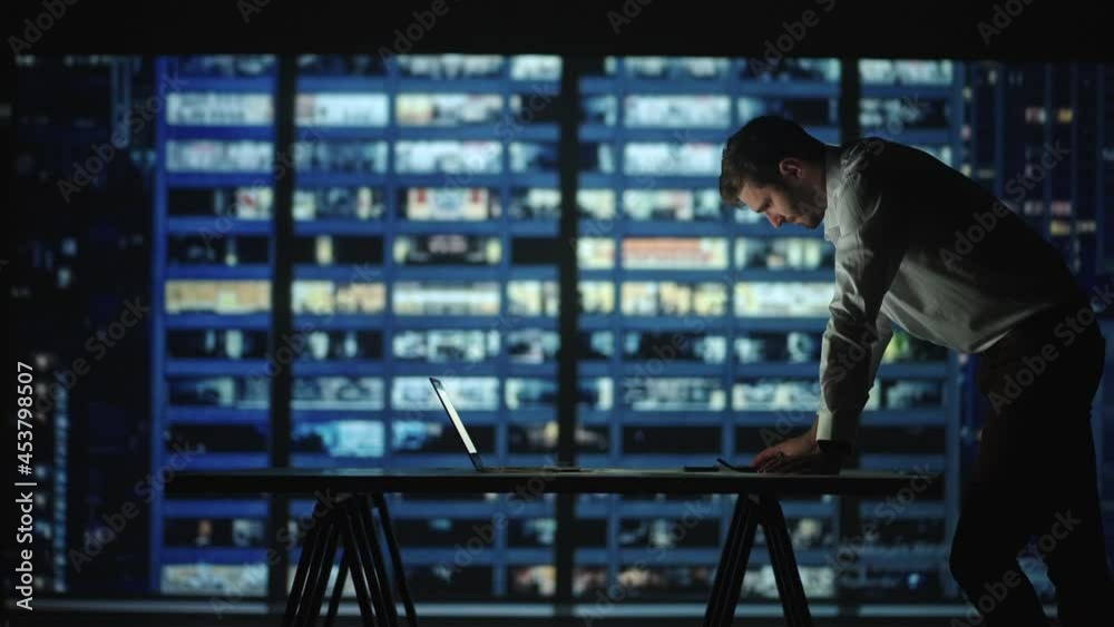 Tired young man working on a laptop late night in the office. Sleepy Businessman sitting at desk in dark office. Tired and stressed businessman in glasses works on a laptop of the night city office
