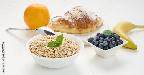 Raw oatmeal in white ceramic plate, blueberries, orange, banana, on white table, breakfast