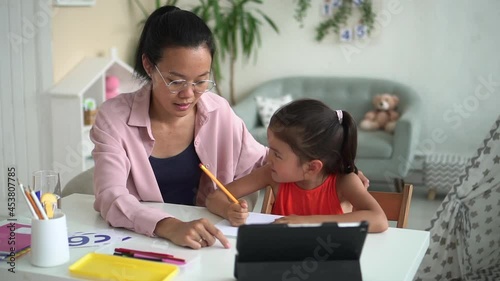 Online primary school education. Spbd Cute little Korean schoolgirl writes in copybook doing hometask with mommy nodding head during video lesson at home photo