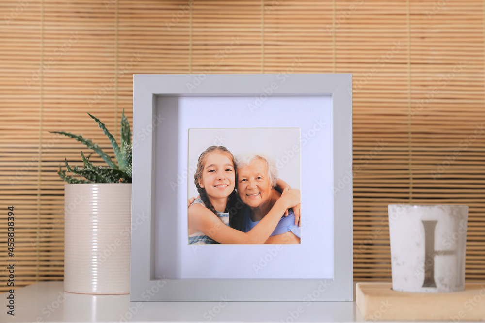 Frame with photo of elderly woman and her granddaughter on white table indoors Frame with photo of elderly woman and her granddaughter on white table indoors
