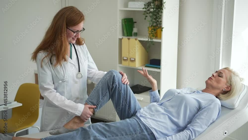 Doctor examines patient in clinic. Spbd Positive lady traumatologist specialist of physical therapy bends leg and touches knee of talking mature woman on daybed Doctor examines patient in clinic. Spbd Positive lady traumatologist specialist of physical therapy bends leg and touches knee of talking mature woman on daybed