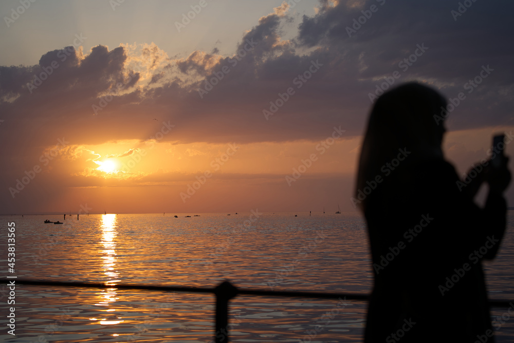 Silhouette of young woman with head scarf taking photo of beautiful sunset at lake Bodensee on sunny summer evening. Photo taken August 15th, 2021, Bregenz, Austria.