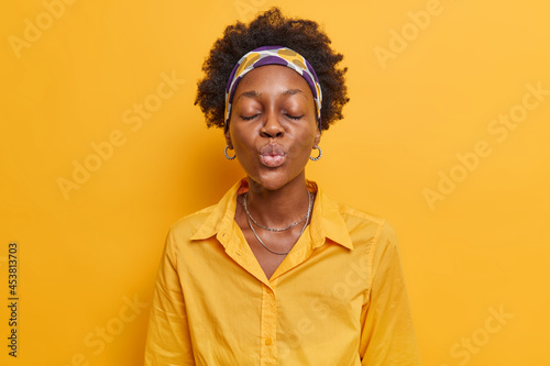 Beautiful dark skinned Afro American woman with puckered lips waits for kiss has romantic mood shows affection wears shirt and headband poses against vivid yellow background sends mwah to you