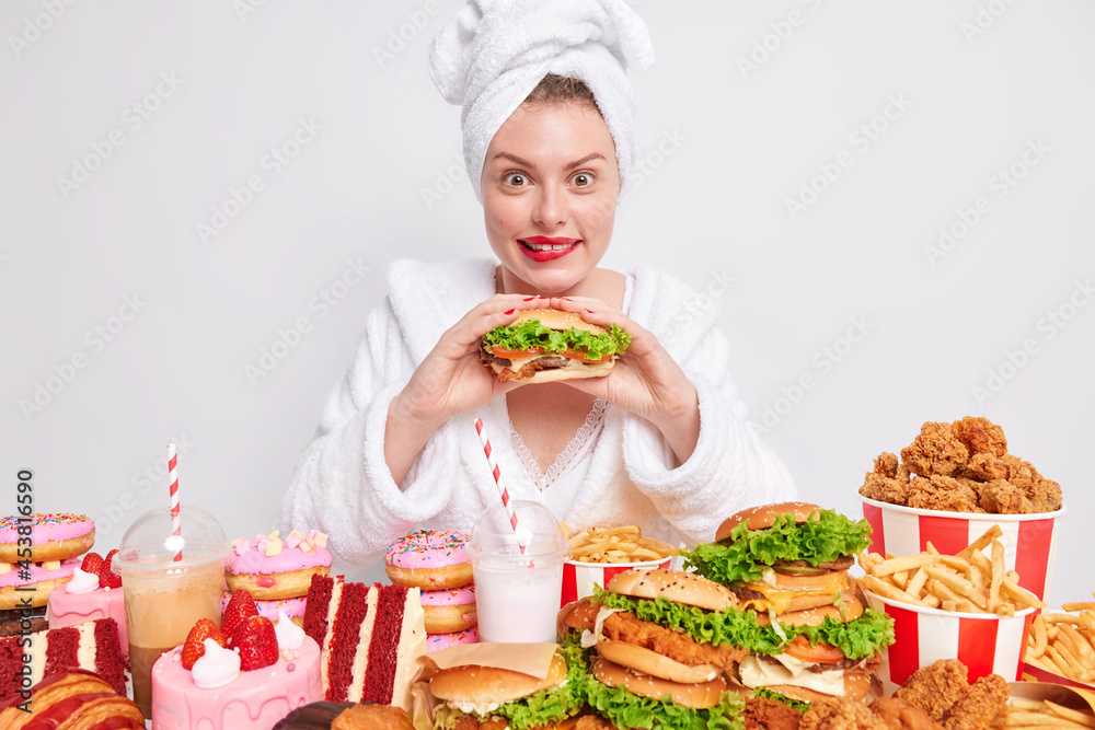 Hungry woman eats greedily burger likes cheat meal and unhealthy junk food has overeating habit wears bathrobe and towel on head surrounded by various tasty treaats against white background.