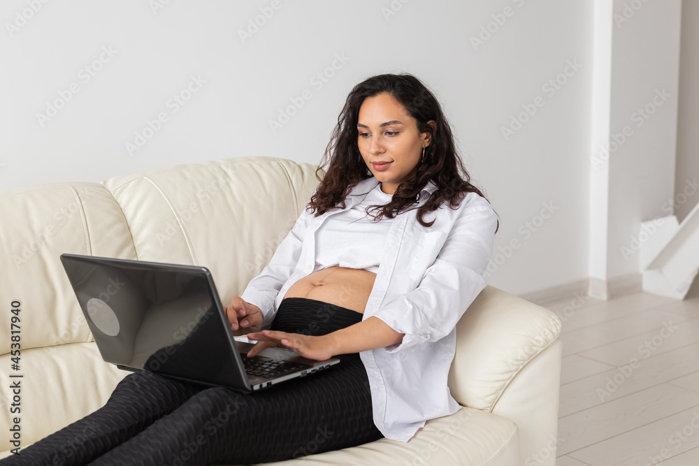 Pregnant woman using laptop while sitting on a couch in the living room at home Pregnant woman using laptop while sitting on a couch in the living room at home