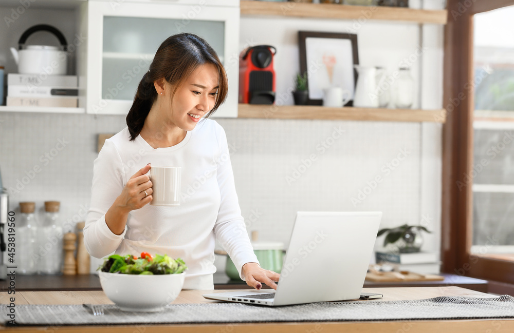 Happy Asian woman working at home using laptop in kitchen in tne morning