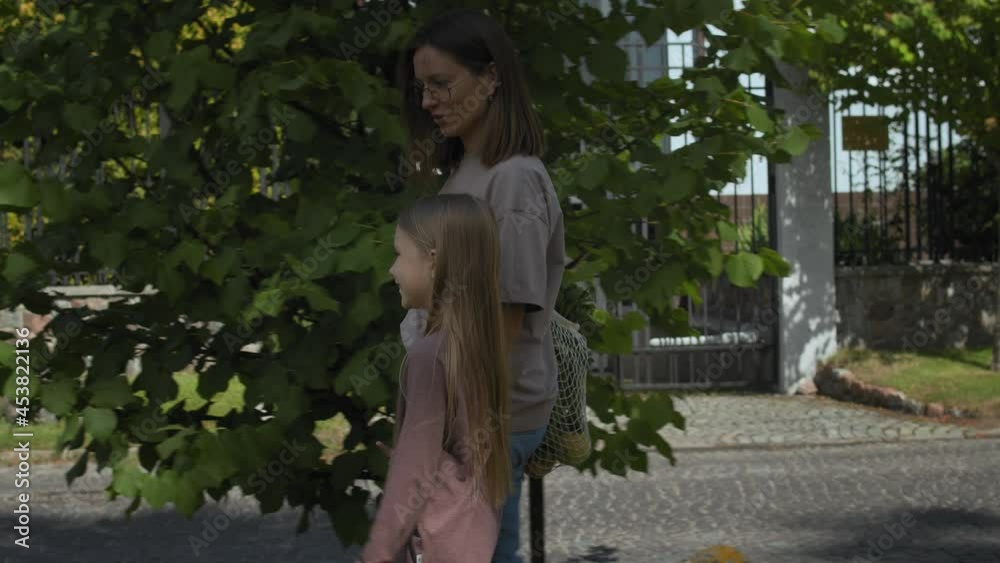 Young woman hold her daughters hand while walking after shopping on city street. She is wearing eco string bag full of groceries. Conscious consumption concept