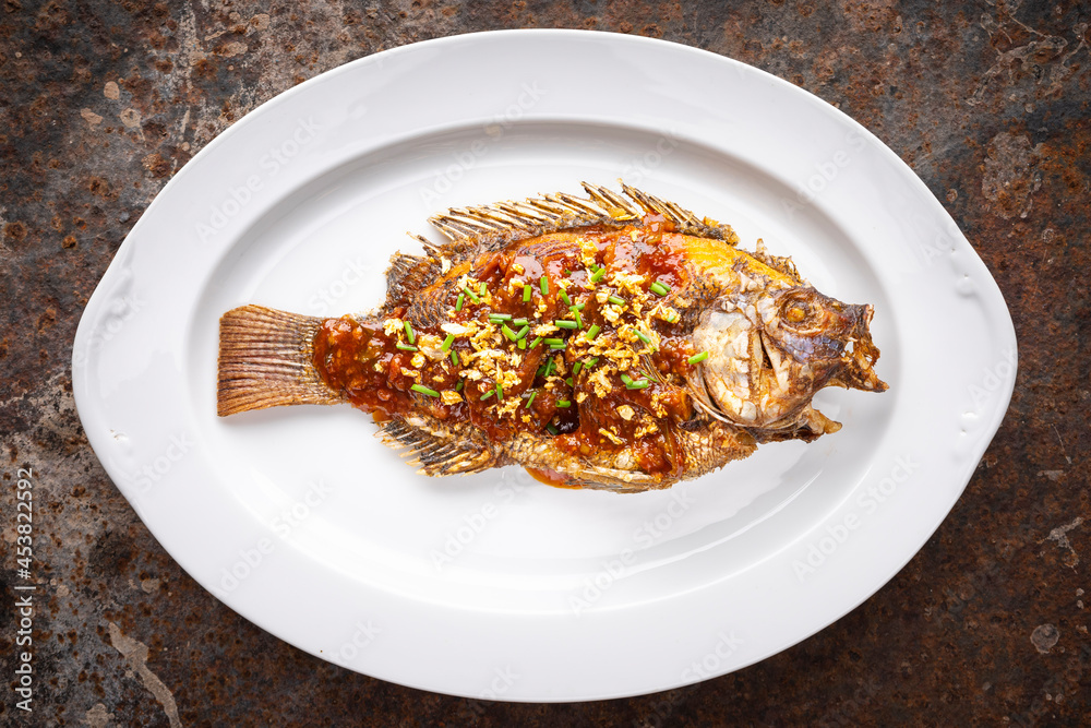 tasty large deedp fried nile tilapia fish with chlli sauce, fried garlic and sliced spring onion in oval ceramic plate on rusty texture background, top view tasty large deedp fried nile tilapia fish with chlli sauce, fried garlic and sliced spring onion in oval ceramic plate on rusty texture background, top view