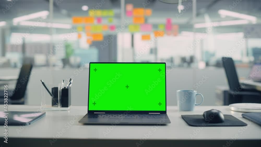 Laptop Computer with Mock Up Green Screen Chroma Key Display Standing on the Desk in the Modern Creative Office. In the Background Glass Wall with Colorful Planning Notes. Zoom in Shot. Laptop Computer with Mock Up Green Screen Chroma Key Display Standing on the Desk in the Modern Creative Office. In the Background Glass Wall with Colorful Planning Notes. Zoom in Shot.