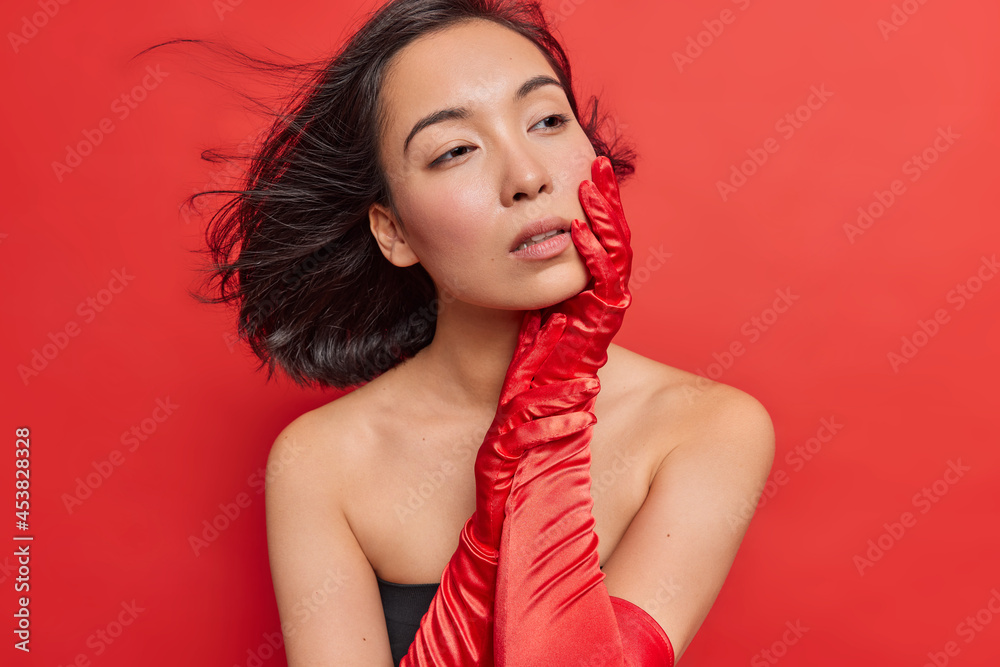 Beautiful Asian woman looks tenderly aside has dark hair floating in air thoughtful expression healthy well cared skin wears long gloves poses against vivid red background. Sexy ethnic female model Beautiful Asian woman looks tenderly aside has dark hair floating in air thoughtful expression healthy well cared skin wears long gloves poses against vivid red background. Sexy ethnic female model