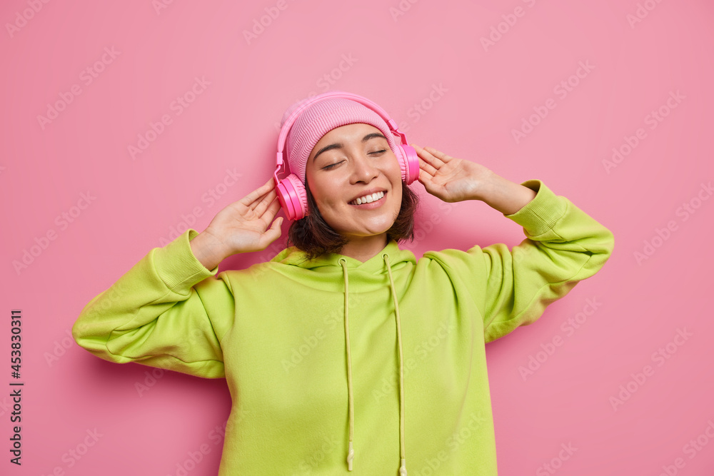 Horizontal shot of happy Asian teenage girl enjoys good sound quality in new headphones listens favorite music closes eyes from satisfaction smiles broadly wears hoodie and hat isolated on pink wall