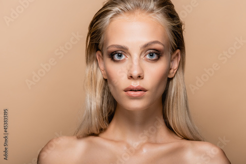 blonde woman with makeup and naked shoulders looking at camera isolated on beige