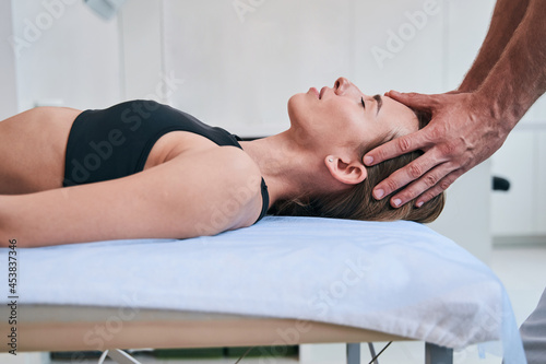 Young elegant Caucasian woman in black sports bra enjoying facial massage in medical office