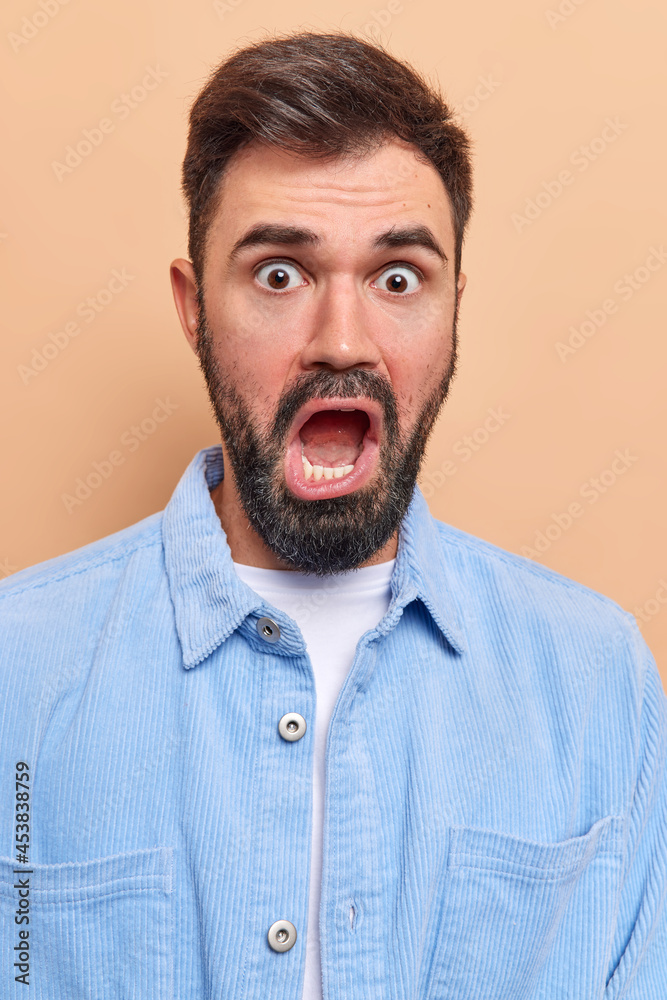 Shocked indignant bearded adult man keeps mouth wide opened looks embarrassed at camera hears unexpected surprising news wears blue velvet shirt stares with indignation. Human reactions concept. Shocked indignant bearded adult man keeps mouth wide opened looks embarrassed at camera hears unexpected surprising news wears blue velvet shirt stares with indignation. Human reactions concept.