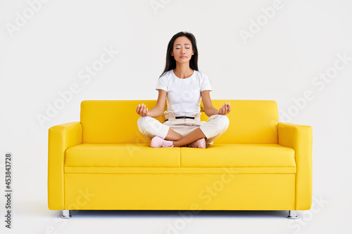Beautiful young asian woman sitting in yoga lotus pose and meditating on comfortable yellow sofa © GVS