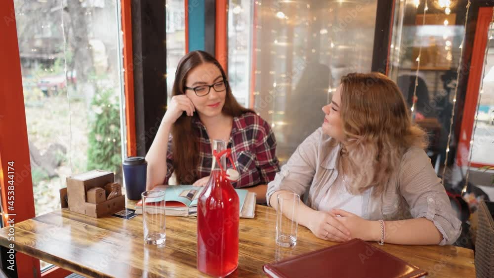 Two pretty female students reading book in a cafe during a coffee break sharing their study notes with lights running on the background reading a books. 4K footage. Two pretty female students reading book in a cafe during a coffee break sharing their study notes with lights running on the background reading a books. 4K footage.