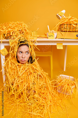 Dispeased female office worker covered with cut paper sits on floor in cabinet has deadline to prepare project work looks unhappily away isolated over yellow background. People job occupation photo