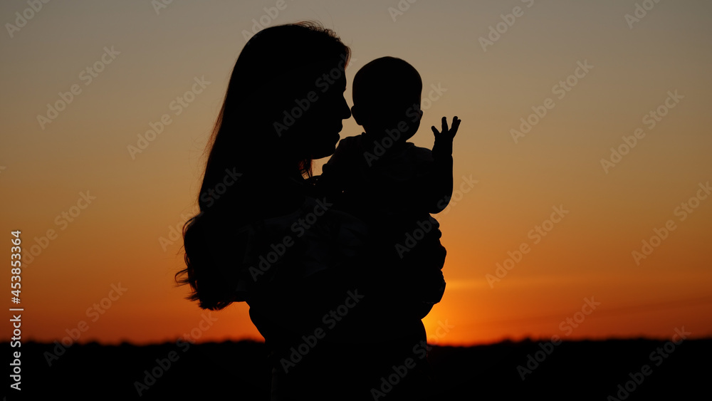 Caring mother holds the baby in her arms and kisses him, unrecognizable silhouette at sunset, motherly love