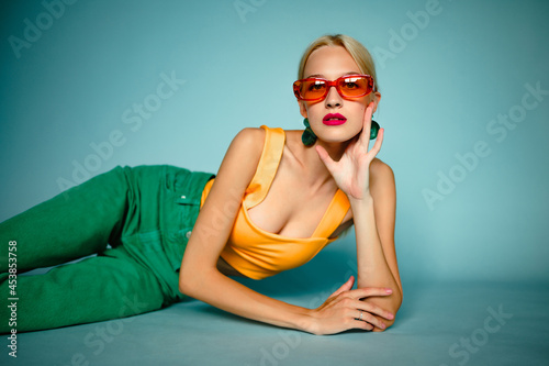 Fashionable confident woman wearing trendy summer orange sunglasses, top, green jeans, posing on blue background. Copy, empty space for text
 photo