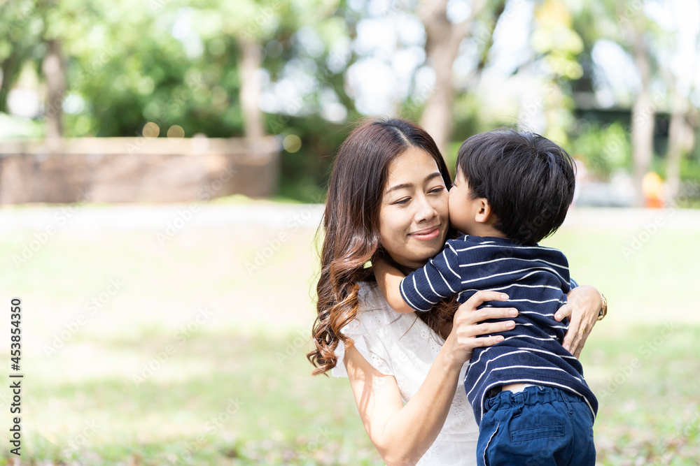 Asian little boy playing and kissing his mother outdoor in the park. Happy mother having fun with her little son outdoor. Young mom spending time with her son on holiday in the garden. people, family