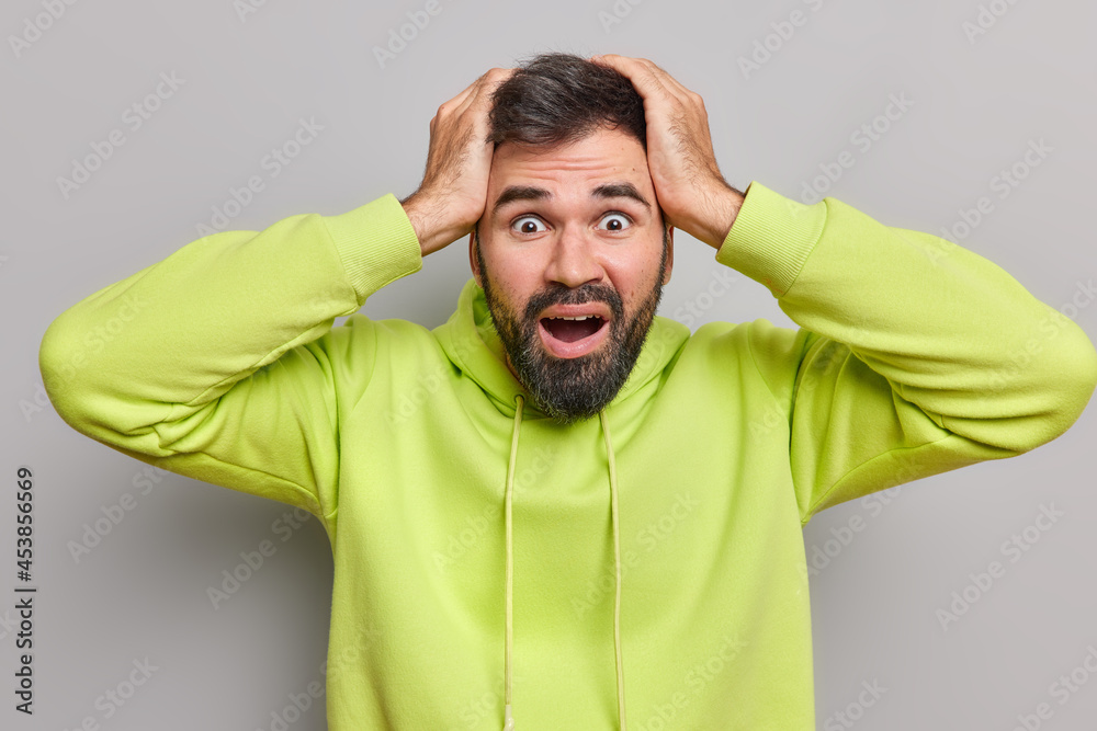Emotional bearded young man touches head with shock has coronavirus panic looks puzzled doesnt know what to do at difficult situation dressed in hoodie poses against grey background has phobia Emotional bearded young man touches head with shock has coronavirus panic looks puzzled doesnt know what to do at difficult situation dressed in hoodie poses against grey background has phobia