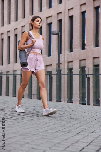 Full length shot of sporty woman in activewear walks in city carries rolled up karemat goes to gym to have fitness training poses against ancient building. People healthy lifestyle and sport