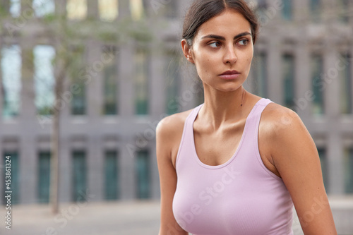 Horizontal shot of healthy thoughtful young sportswoman in casual t shirt looks away pensively walks outdoor during daytime poses against blurred building background notices something on street