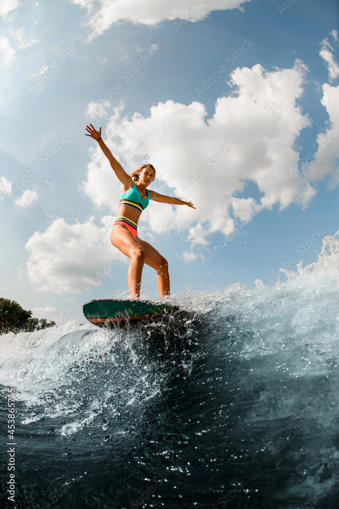 young woman having fun wakesurfing on the board on the river wave against blue sky