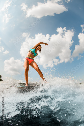 Happy smiling young woman in colorful swimsuit energetically balancing on wave on wakesurf board.
