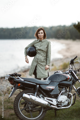 Portrait of a female biker standing outdoors. A woman holds a helmet in her hands on the background of a motorcycle.