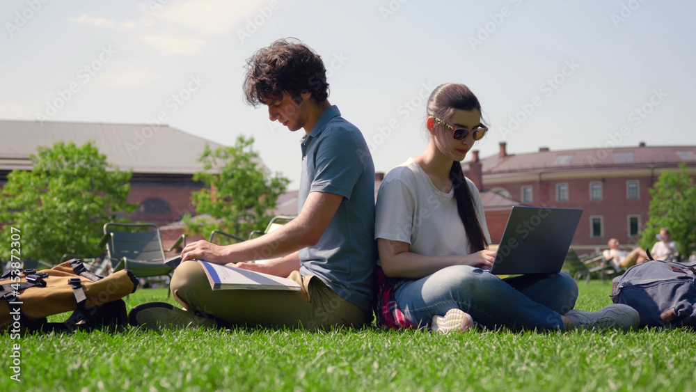 Couple of students sitting in park and studying with laptop and textbook