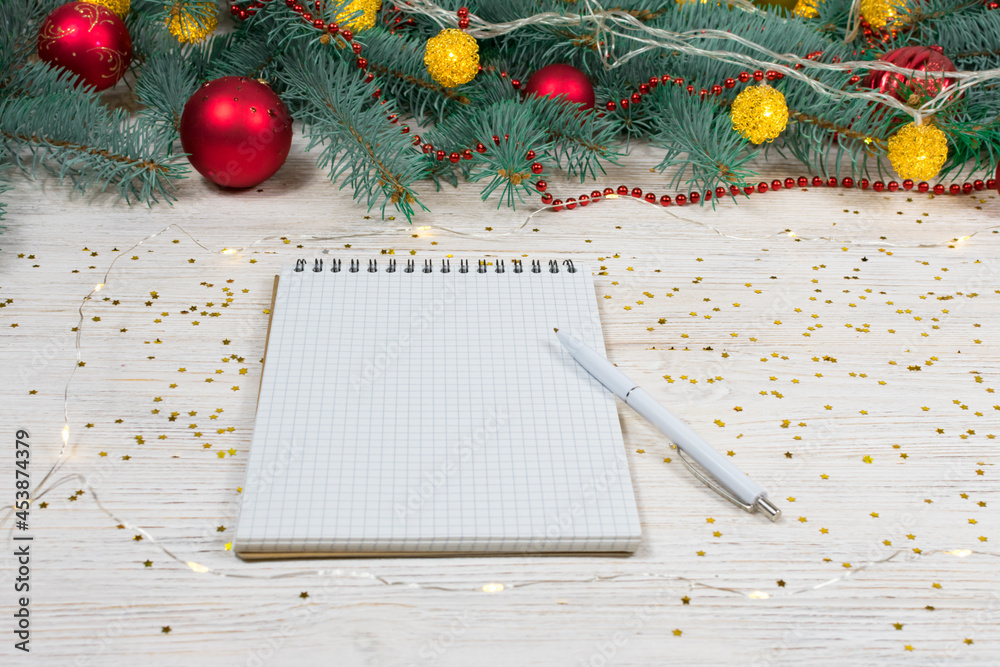 Blank notebook with a white pen on a white table with gold sparkles, stars on the background of a Christmas tree decorated with toys. Festive concept. Place for an inscription. Blank notebook with a white pen on a white table with gold sparkles, stars on the background of a Christmas tree decorated with toys. Festive concept. Place for an inscription.