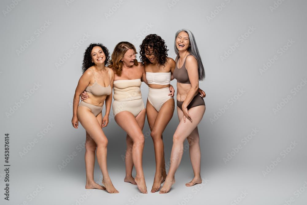 Smiling body positive women embracing multiethnic friends on grey background