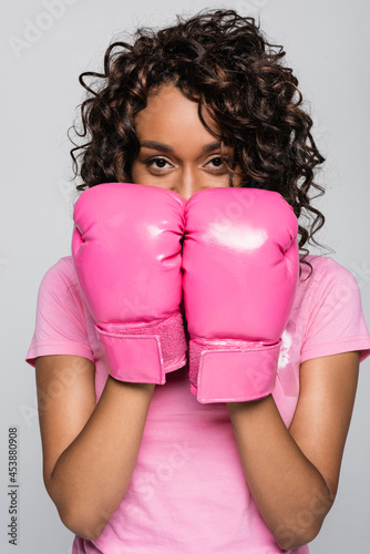 African american woman with pink ribbon and boxing gloves looking at camera isolated on grey