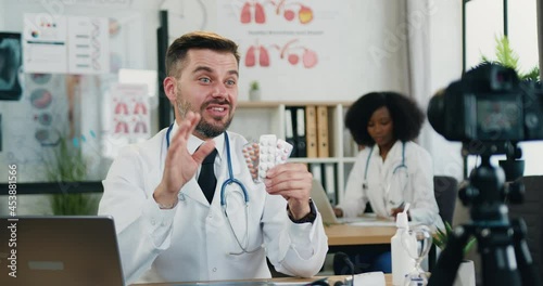 Good-looking confident skilled adult bearded doctor holding packs of pills in his hand during recording videovlog for medical channel on professional camera photo