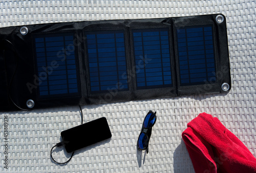 Top view of portable solar panel charging cell phone next to sunglasses and red towel on a white deck chair