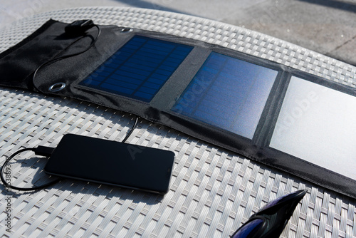 Portable solar panel charging cell phone next to sunglasses on a white deck chair