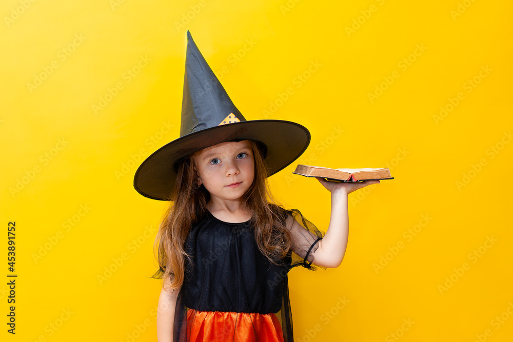 Halloween. A serious cute girl in a witch costume holds an old book in her hand. Colored Yellow background. A place for text. Halloween. A serious cute girl in a witch costume holds an old book in her hand. Colored Yellow background. A place for text.