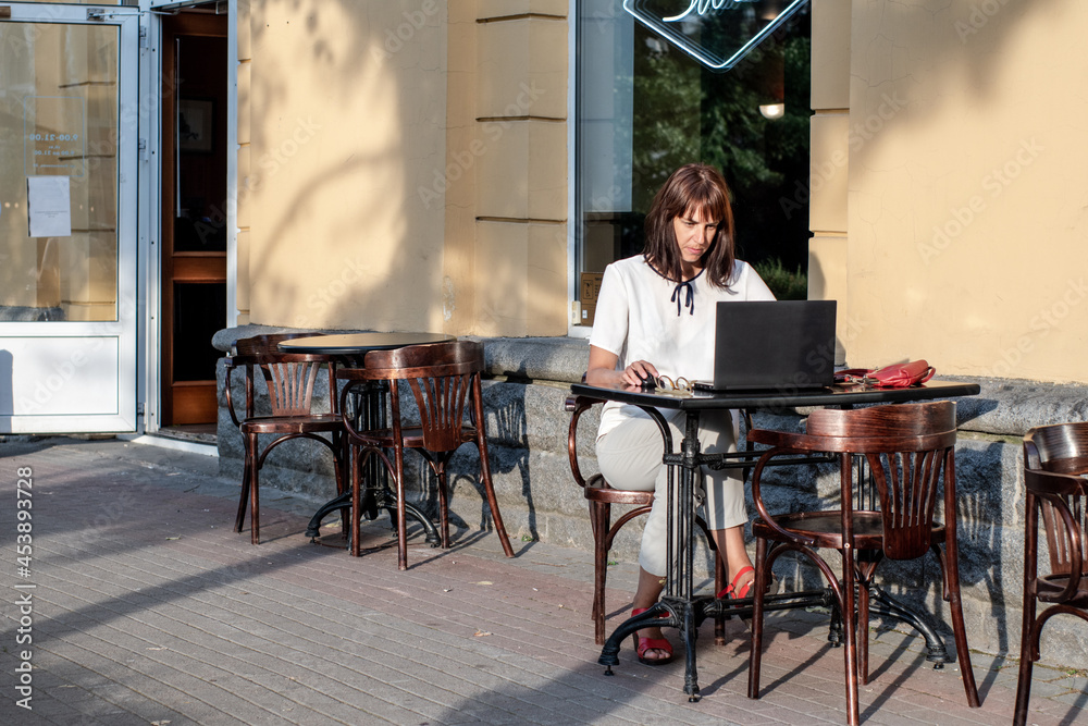 Fototapeta premium A young woman is sitting at a laptop in a summer street cafe. people using social media