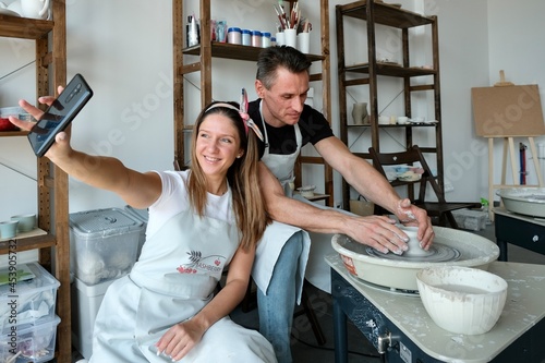 A young couple takes pictures of themselves on a smartphone in a pottery studio. A young couple work on a potter's wheel. Creativity, hobby, privacy. Couple Using  smartphone