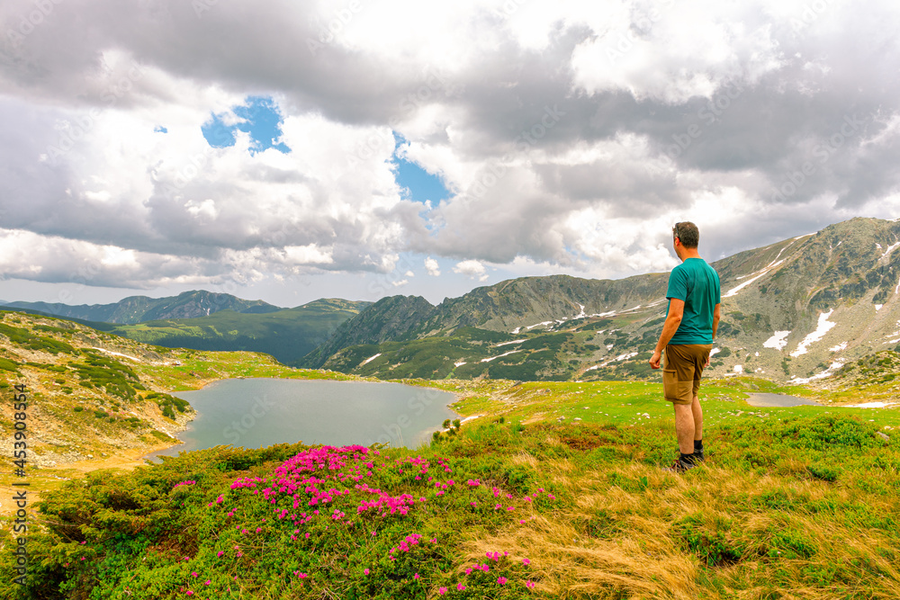 Photography of a male hiker in the mountains admiring a beautiful lake down in the valley in summer time with cloudy weather.