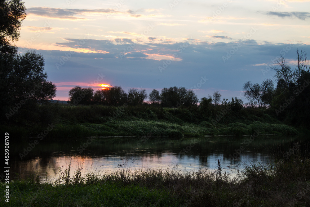 A beautiful sunrise over the river and black silhouettes of trees against the background of a cloudy dramatic sky in the early summer morning and a space to copy