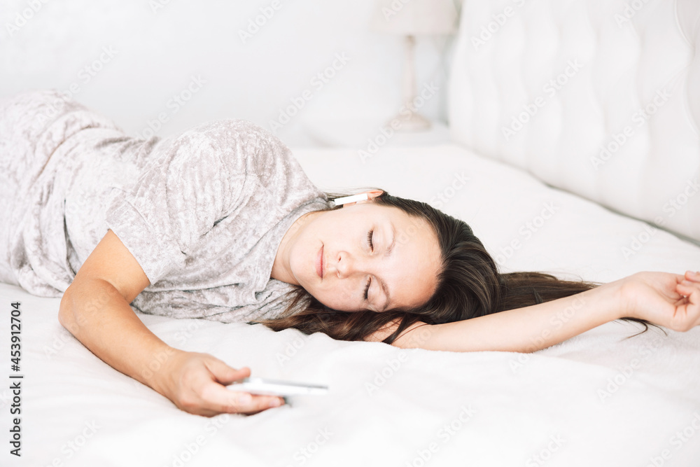 Beautiful brunette girl is lying on bed with wireless headphones and white smartphone in her hands and listening to music or audiobook. Young woman with smartphone sleeping in bed at bedroom Beautiful brunette girl is lying on bed with wireless headphones and white smartphone in her hands and listening to music or audiobook. Young woman with smartphone sleeping in bed at bedroom