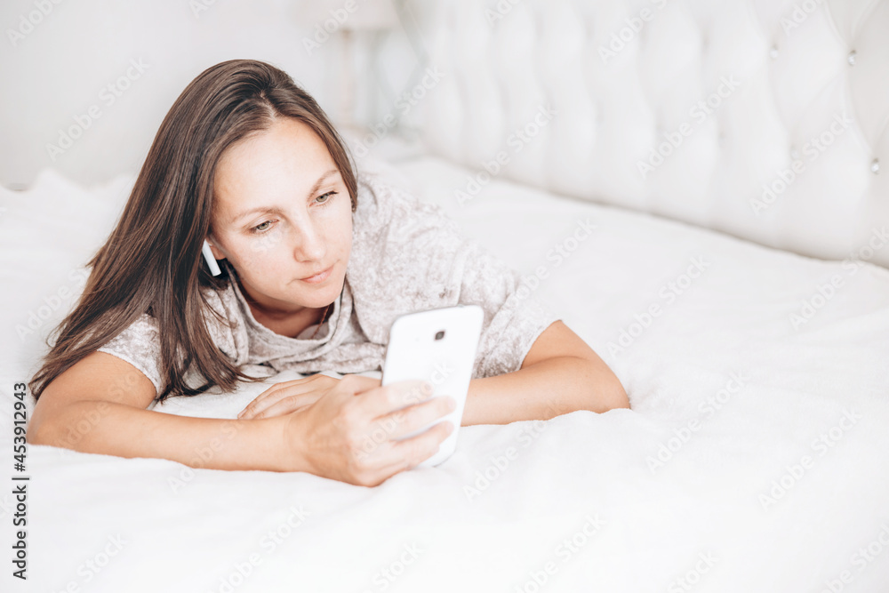 Beautiful brunette girl is lying on bed with wireless headphones and white smartphone in her hands and listening to music or audiobook. Beautiful brunette girl is lying on bed with wireless headphones and white smartphone in her hands and listening to music or audiobook.