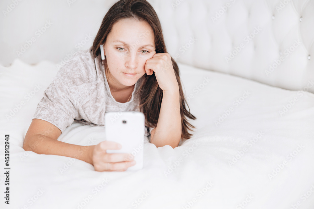 Beautiful brunette girl is lying on bed with wireless headphones and white smartphone in her hands and listening to music or audiobook. Beautiful brunette girl is lying on bed with wireless headphones and white smartphone in her hands and listening to music or audiobook.